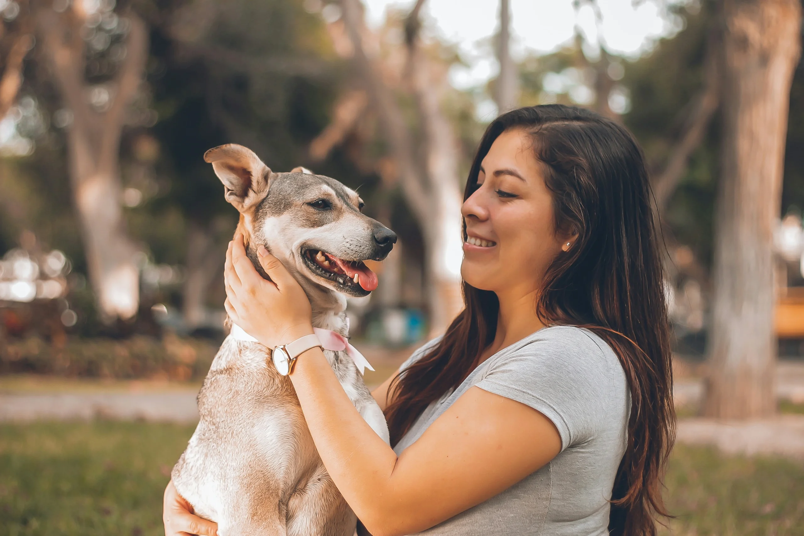 A smiling young woman gently holding and looking affectionately at her happy, panting dog in a sunlit park.